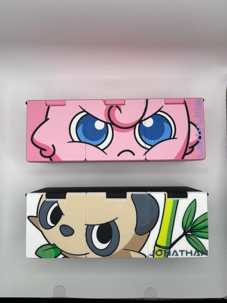 May include: Two colorful, hinged boxes. The top box is pink with a cartoon face and blue eyes. The bottom box is white and tan with a cartoon face, green bamboo, and the name "JONATHAN".