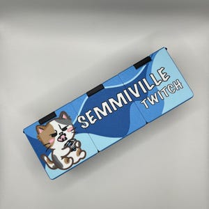 Może przedstawiać: Niebiesko-biały prostokątny przedmiot z napisem "SEMMIVILLE TWITCH" i ilustracją kota z kresk&oacute;wki. Kot jest brązowy, biały i szary, trzymający mały przedmiot. Przedmiot ma czarne zawiasy.