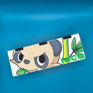 May include: A white, rectangular, folding item featuring a cartoon panda-like character with a stern expression. The design includes green leaves, vertical green and yellow stripes, and the name "NICO" in green letters. The item is set against a blue background.