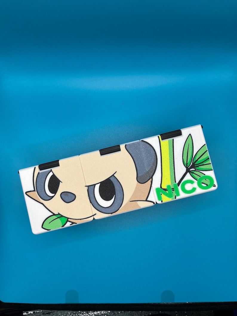 May include: A white, rectangular, folding item featuring a cartoon panda-like character with a stern expression. The design includes green leaves, vertical green and yellow stripes, and the name "NICO" in green letters. The item is set against a blue background.