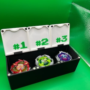 May include: A black storage box with three numbered compartments holding colorful spinning tops. The compartments are labeled #1, #2, and #3 in green. The tops are red, green, and purple.