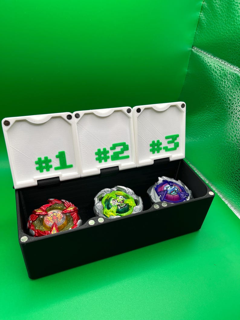 May include: A black storage box with three numbered compartments holding colorful spinning tops. The compartments are labeled #1, #2, and #3 in green. The tops are red, green, and purple.