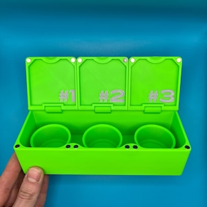 May include: A vibrant green container with three compartments and numbered lids. Inside are three matching green cups. The background is a solid blue. The container is held by a hand.