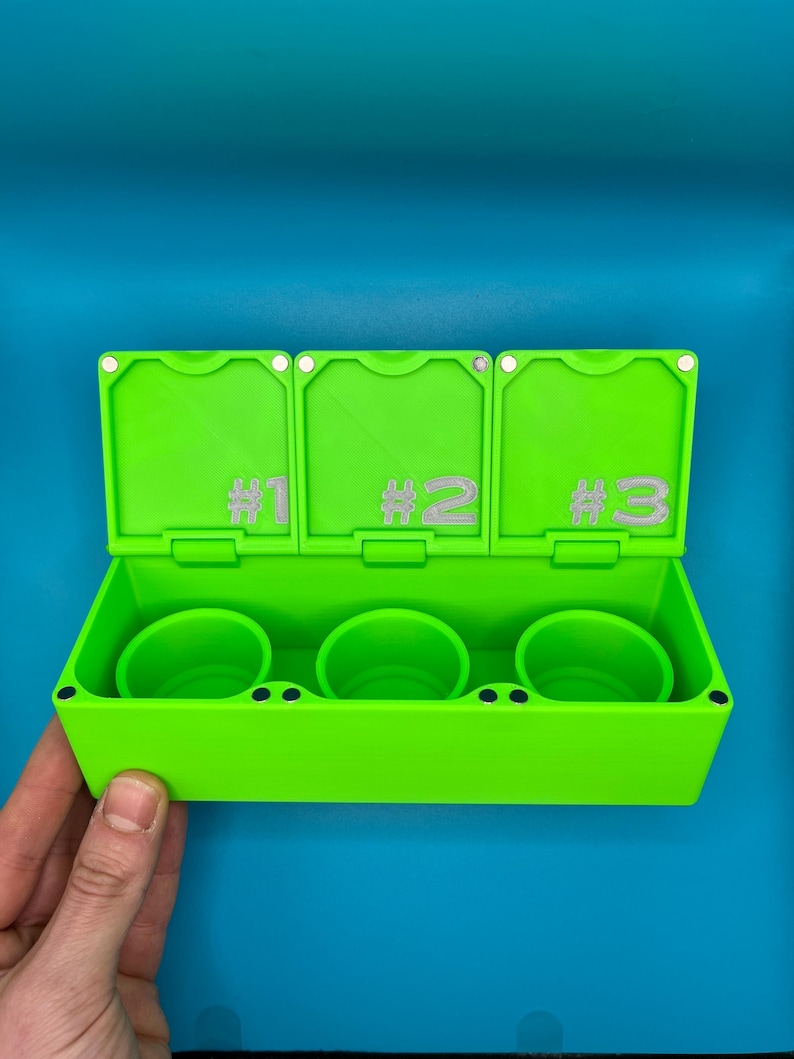 May include: A vibrant green container with three compartments and numbered lids. Inside are three matching green cups. The background is a solid blue. The container is held by a hand.
