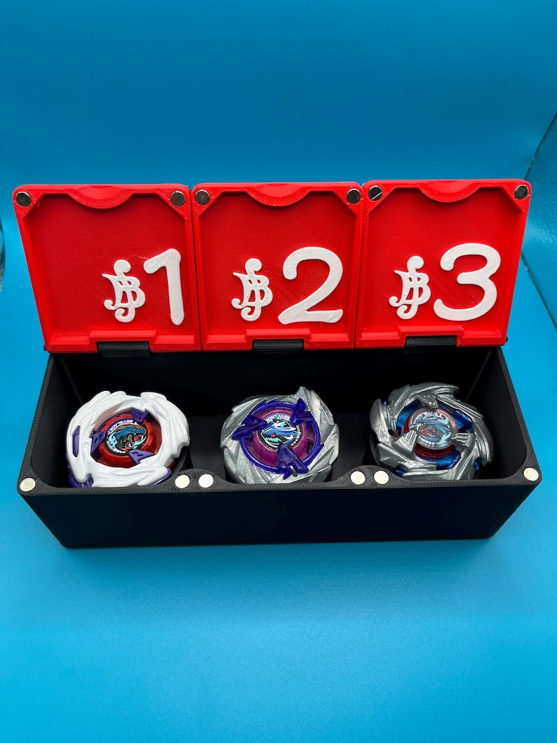 Peut inclure: Une bo&icirc;te de rangement noire avec trois couvercles rouges &agrave; charni&egrave;res, chacun portant un num&eacute;ro blanc et un symbole mon&eacute;taire. &Agrave; l'int&eacute;rieur, trois toupies color&eacute;es. Le fond est bleu uni.