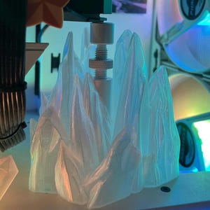 Op de afbeelding: Een wit 3D-geprint model van een bergketen, met een getextureerd oppervlak, wordt geprint op een 3D-printer. Het model wordt op zijn plaats gehouden door een metalen staaf.