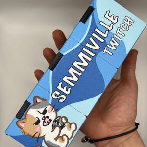 Może przedstawiać: Niebiesko-biała zabawka typu fidget z napisem "SEMMIVILLE TWITCH" i ilustracją kota z kresk&oacute;wek. Zabawka, trzymana w dłoni, ma czarne elementy łączące.