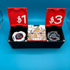 Peut inclure: Une bo&icirc;te noire avec des couvercles rouges marqu&eacute;s &laquo;&nbsp;$1&nbsp;&raquo; et &laquo;&nbsp;$3&nbsp;&raquo; contient deux toupies et une image de personnage. Les toupies sont blanches et argent&eacute;es avec des motifs color&eacute;s. L'image du personnage pr&eacute;sente un visage souriant avec des cheveux blancs et des accents rouges.