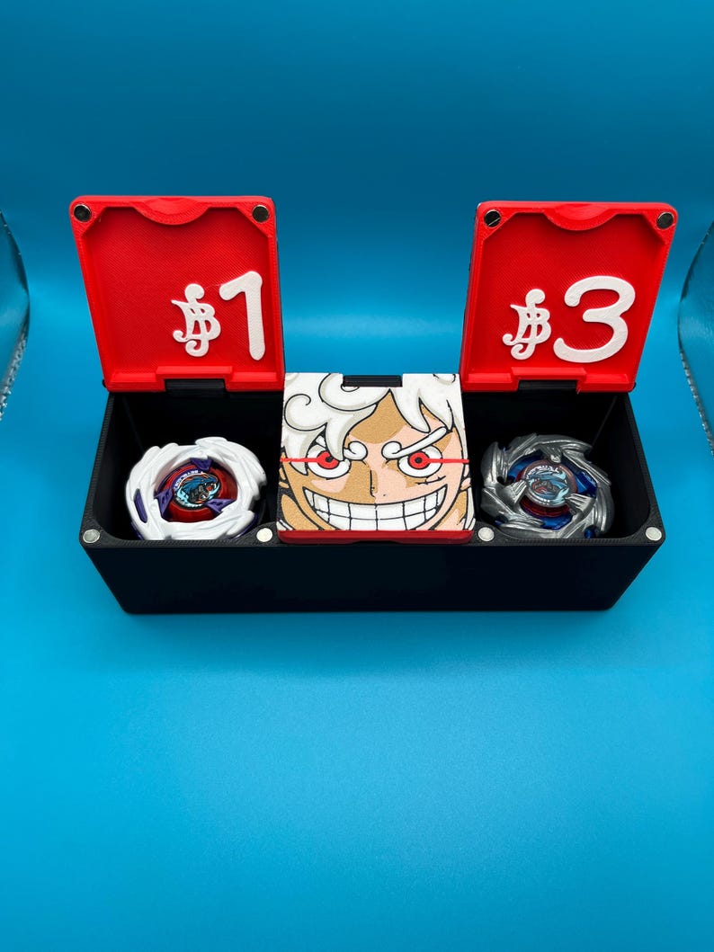 Peut inclure: Une bo&icirc;te noire avec des couvercles rouges marqu&eacute;s &laquo;&nbsp;$1&nbsp;&raquo; et &laquo;&nbsp;$3&nbsp;&raquo; contient deux toupies et une image de personnage. Les toupies sont blanches et argent&eacute;es avec des motifs color&eacute;s. L'image du personnage pr&eacute;sente un visage souriant avec des cheveux blancs et des accents rouges.