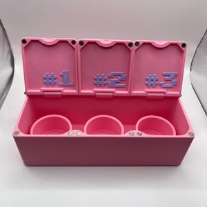 May include: A pink storage box with three compartments, each with a circular holder. The box has three hinged lids, each labeled with a number in purple: #1, #2, and #3. The box is open, revealing the interior.