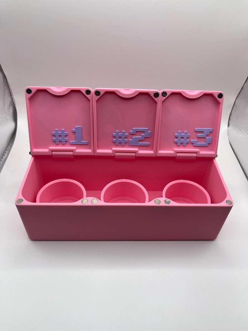 May include: A pink storage box with three compartments, each with a circular holder. The box has three hinged lids, each labeled with a number in purple: #1, #2, and #3. The box is open, revealing the interior.