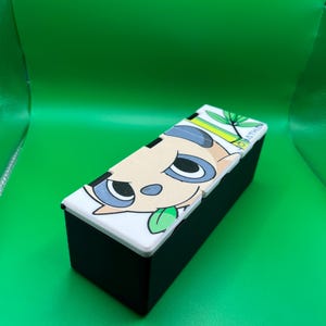 May include: A black rectangular box with a white lid featuring a cartoon animal face with blue eyes and a green tongue. The box is set against a bright green background.