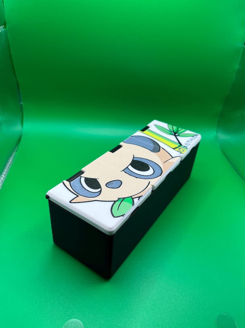 May include: A black rectangular box with a white lid featuring a cartoon animal face with blue eyes and a green tongue. The box is set against a bright green background.