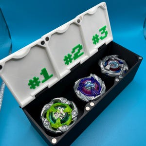 May include: A black storage box with a white lid, holding three colorful spinning tops. The lid is labeled with green numbers #1, #2, and #3. The tops have various colors, including green, blue, and purple, and are designed with intricate details.