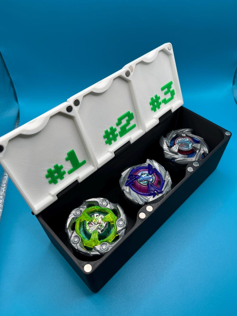 May include: A black storage box with a white lid, holding three colorful spinning tops. The lid is labeled with green numbers #1, #2, and #3. The tops have various colors, including green, blue, and purple, and are designed with intricate details.