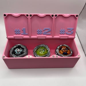 May include: A pink storage box with three compartments, each labeled with a number. The box contains three spinning tops in various colors. The tops are silver, green, and orange.