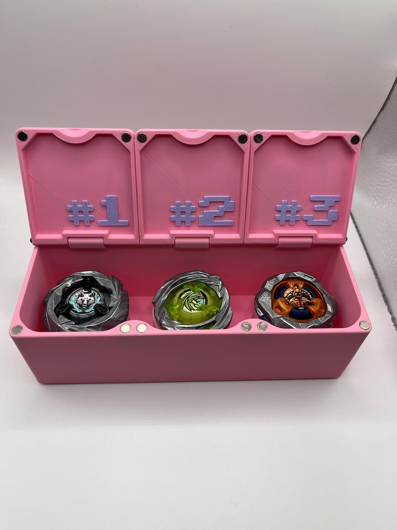 May include: A pink storage box with three compartments, each labeled with a number. The box contains three spinning tops in various colors. The tops are silver, green, and orange.
