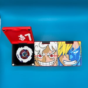 Peut inclure: Une bo&icirc;te de rangement de jouets rouge et noire avec un toupie blanche et violette &agrave; l'int&eacute;rieur. L'&eacute;tui pr&eacute;sente des illustrations de personnages de dessins anim&eacute;s et du texte japonais sur un fond bleu.