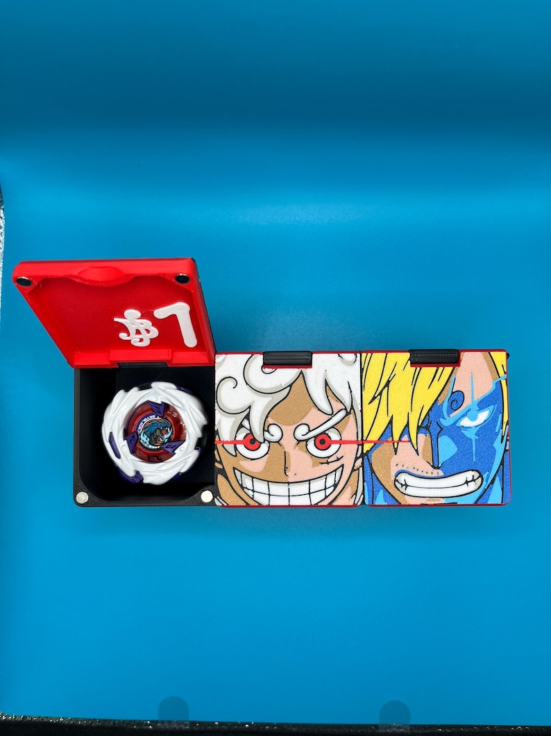 Peut inclure: Une bo&icirc;te de rangement de jouets rouge et noire avec un toupie blanche et violette &agrave; l'int&eacute;rieur. L'&eacute;tui pr&eacute;sente des illustrations de personnages de dessins anim&eacute;s et du texte japonais sur un fond bleu.