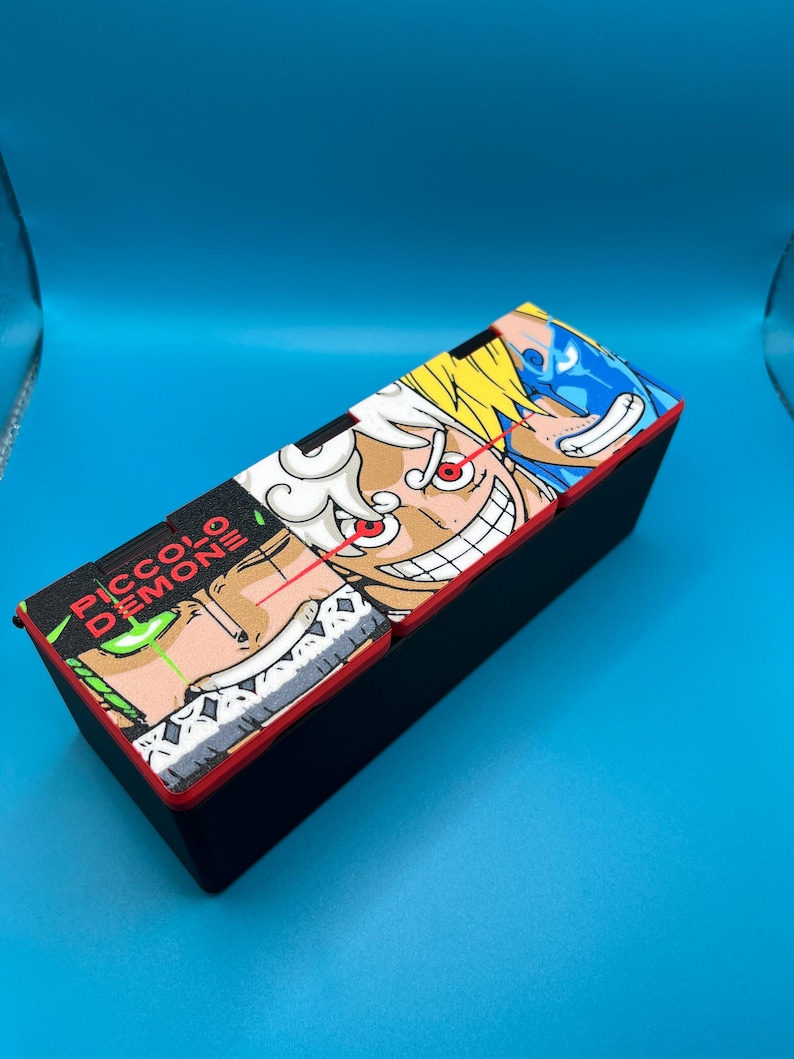 Peut inclure: Bo&icirc;te rectangulaire noire avec une bordure rouge et un graphisme de dessin anim&eacute; color&eacute; sur le couvercle. Le graphisme pr&eacute;sente plusieurs personnages de style anime avec des contours audacieux et des couleurs vives. Le texte "PICCOLO DEMON" est &eacute;galement visible.