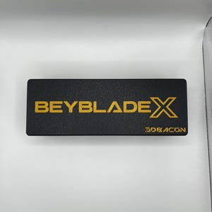 Puede incluir: Una caja rectangular negra con las palabras "BEYBLADE X" en letras doradas. La palabra "3DRAGON" est&aacute; impresa en dorado en la esquina inferior derecha de la caja.