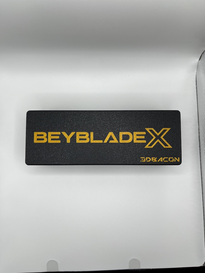 Puede incluir: Una caja rectangular negra con las palabras "BEYBLADE X" en letras doradas. La palabra "3DRAGON" est&aacute; impresa en dorado en la esquina inferior derecha de la caja.
