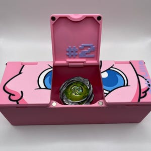 May include: A pink storage box with a hinged lid, featuring a cartoon character design. The box is open, revealing a metallic green and silver spinning top toy. The lid displays the number "#2" in purple.