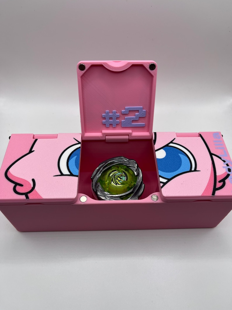 May include: A pink storage box with a hinged lid, featuring a cartoon character design. The box is open, revealing a metallic green and silver spinning top toy. The lid displays the number "#2" in purple.