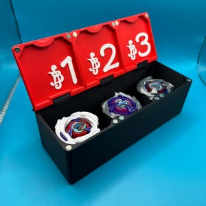Peut inclure: Une bo&icirc;te de rangement noire avec un couvercle rouge, comportant trois compartiments. Le couvercle est num&eacute;rot&eacute; 1, 2 et 3, chacun pr&eacute;c&eacute;d&eacute; d'un symbole dollar. &Agrave; l'int&eacute;rieur, trois toupies color&eacute;es. La bo&icirc;te est pos&eacute;e sur un fond bleu.