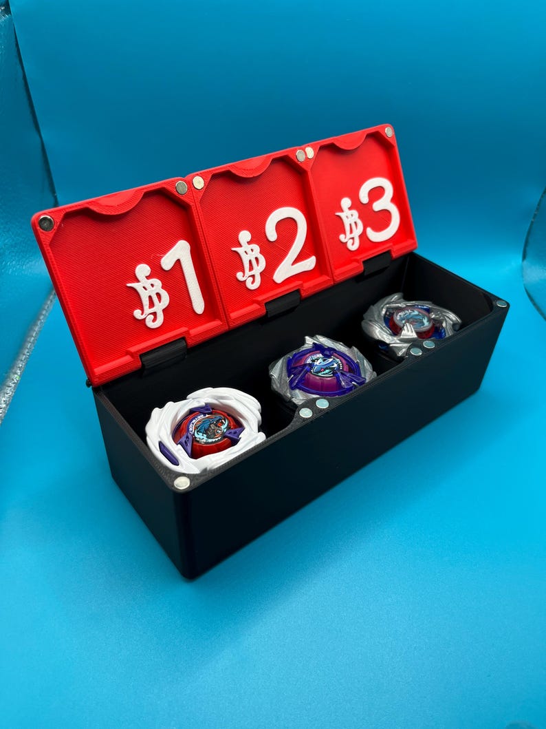 Peut inclure: Une bo&icirc;te de rangement noire avec un couvercle rouge, comportant trois compartiments. Le couvercle est num&eacute;rot&eacute; 1, 2 et 3, chacun pr&eacute;c&eacute;d&eacute; d'un symbole dollar. &Agrave; l'int&eacute;rieur, trois toupies color&eacute;es. La bo&icirc;te est pos&eacute;e sur un fond bleu.