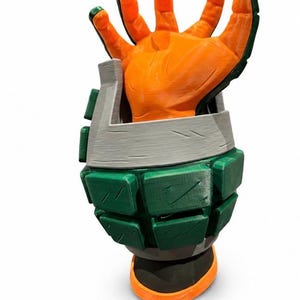 Puede incluir: Un guante de utiler&iacute;a impreso en 3D con una mano naranja de dedos articulados que sobresale de un gran guantelete segmentado en forma de granada verde y gris. El guantelete tiene una base gris oscuro y una superficie texturizada visible.