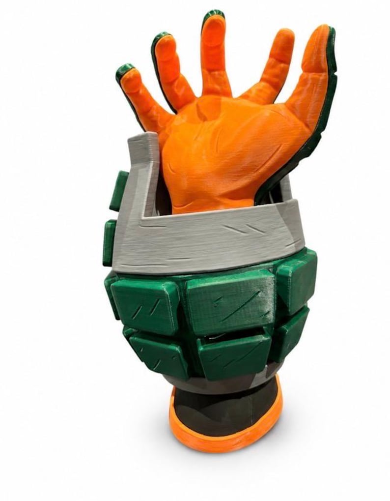 Puede incluir: Un guante de utiler&iacute;a impreso en 3D con una mano naranja de dedos articulados que sobresale de un gran guantelete segmentado en forma de granada verde y gris. El guantelete tiene una base gris oscuro y una superficie texturizada visible.