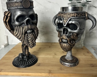 Puede incluir: Dos copas de calavera plateadas con detalles dorados y barbas. Las copas están sobre una superficie de madera.