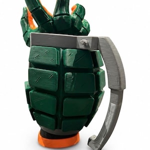 Puede incluir: Una taza verde y naranja con forma de granada de mano, con un cuerpo texturizado y segmentado y un asa gris. La parte superior de la taza presenta una mano esculpida con los dedos extendidos hacia arriba, mientras que la base es un anillo naranja s&oacute;lido.