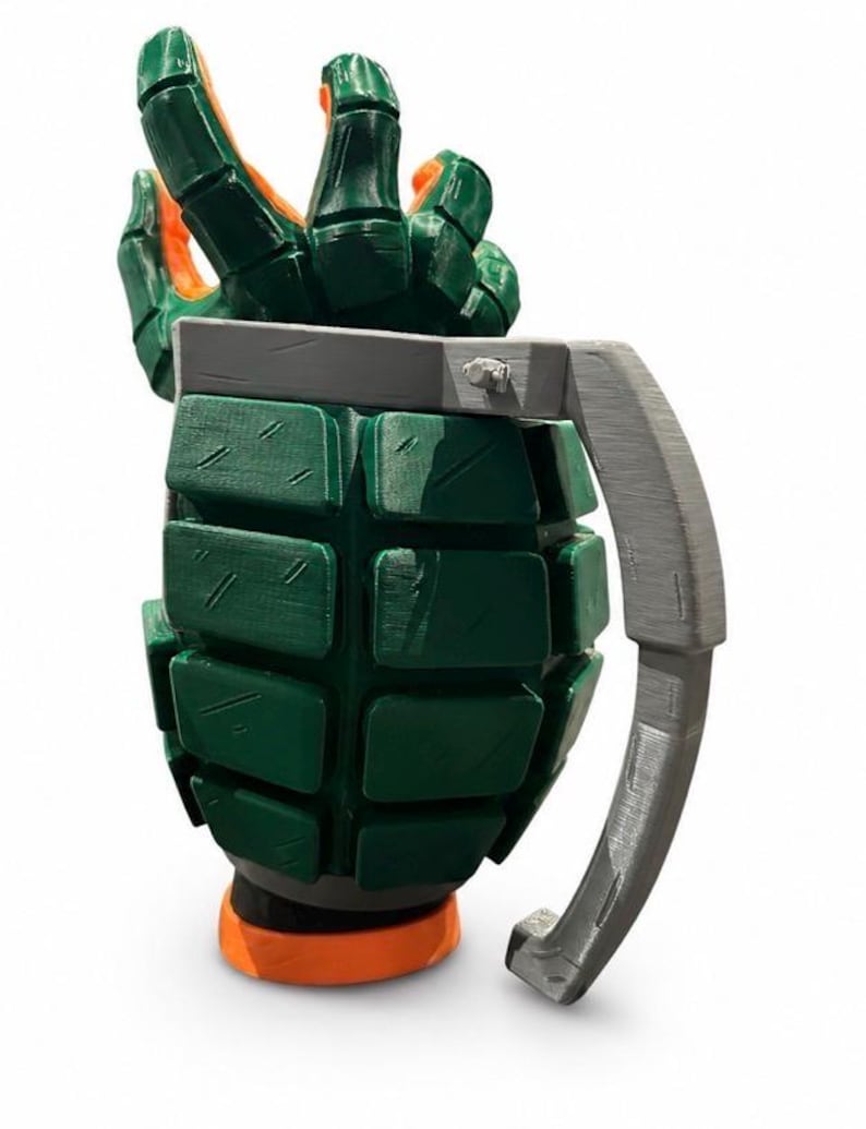 Puede incluir: Una taza verde y naranja con forma de granada de mano, con un cuerpo texturizado y segmentado y un asa gris. La parte superior de la taza presenta una mano esculpida con los dedos extendidos hacia arriba, mientras que la base es un anillo naranja s&oacute;lido.