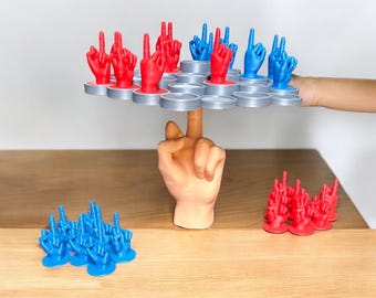 May include: A hand balancing a circular game board with red and blue hand figurines on top. The board is made of gray plastic and has a honeycomb pattern. There are more red and blue hand figurines on the table below the board.