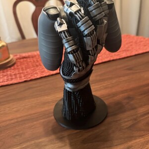 Puede incluir: Un soporte para mando en forma de mano rob&oacute;tica con base negra y antebrazo vertical texturizado en negro. La mano est&aacute; construida con articulaciones y dedos mec&aacute;nicos en color plata met&aacute;lica, dise&ntilde;ados para sostener un mando gris.