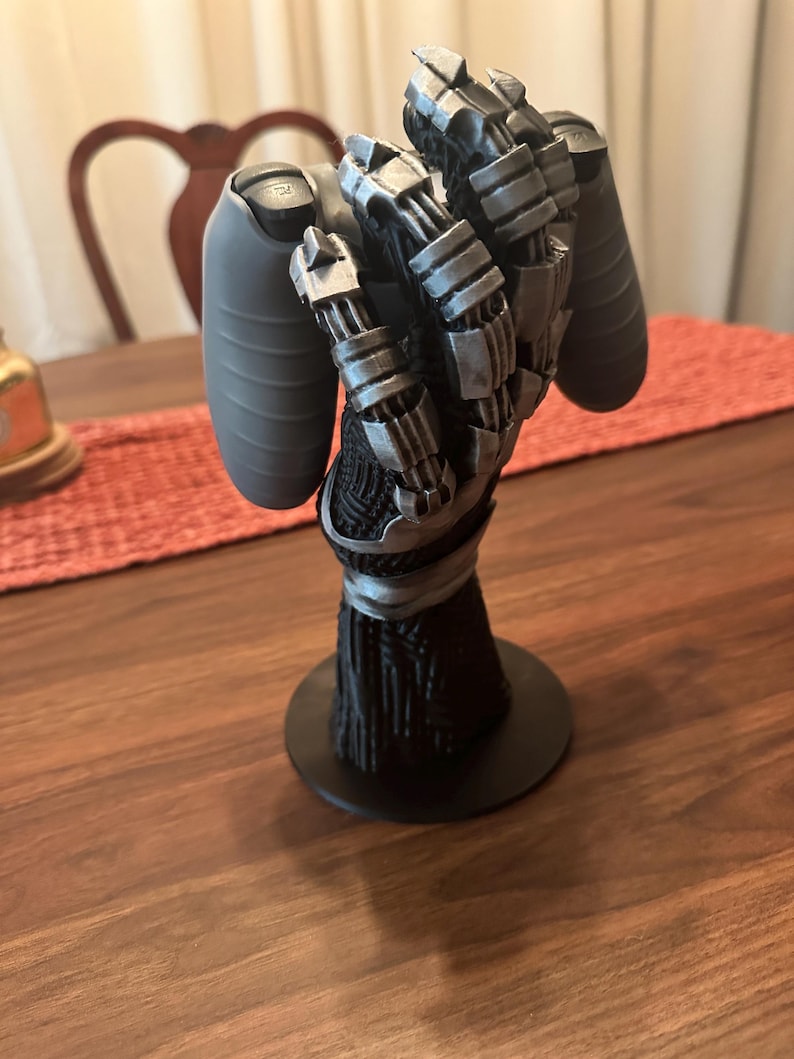 Puede incluir: Un soporte para mando en forma de mano rob&oacute;tica con base negra y antebrazo vertical texturizado en negro. La mano est&aacute; construida con articulaciones y dedos mec&aacute;nicos en color plata met&aacute;lica, dise&ntilde;ados para sostener un mando gris.