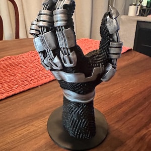 Puede incluir: Un soporte de exhibici&oacute;n con forma de mano esculpida que presenta una base negra texturizada con patrones geom&eacute;tricos intrincados y detalles en plata met&aacute;lica en los dedos y la mu&ntilde;eca. La mano est&aacute; montada sobre una base circular de color negro.