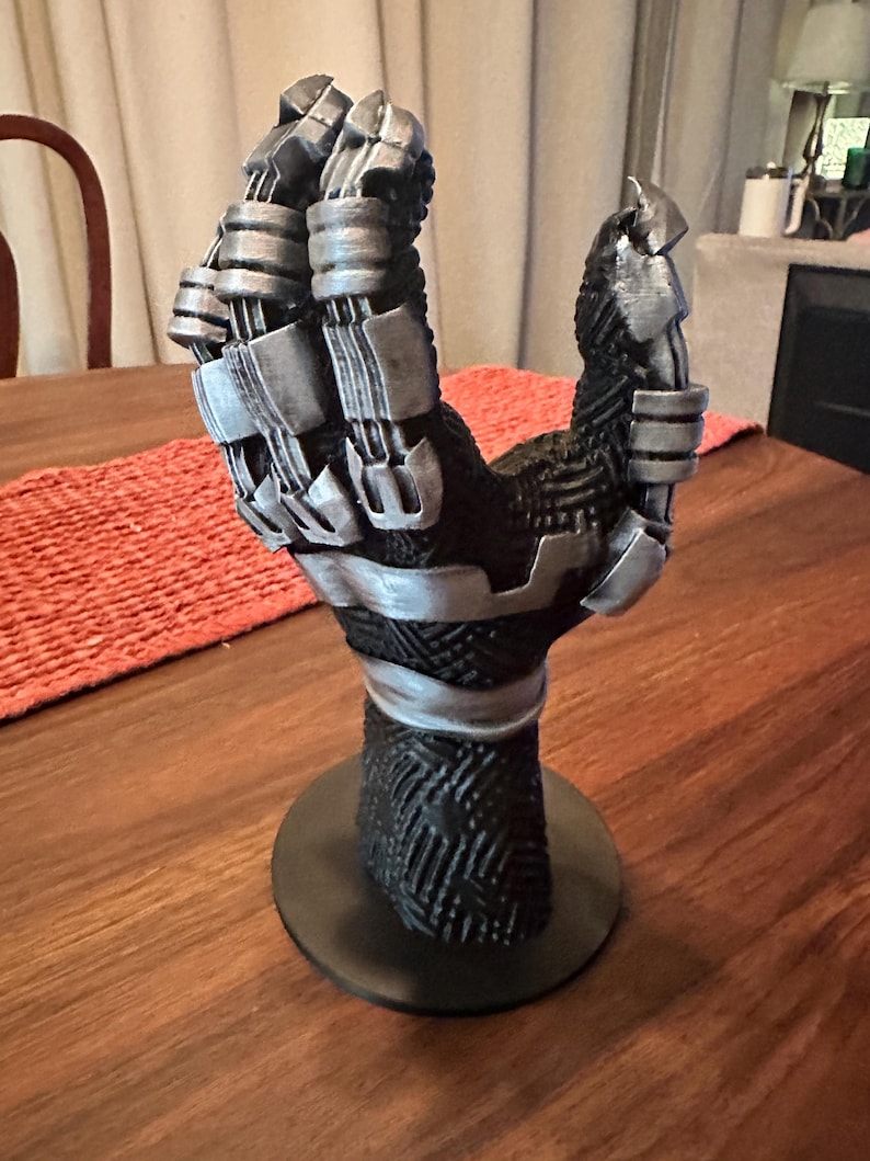 Puede incluir: Un soporte de exhibici&oacute;n con forma de mano esculpida que presenta una base negra texturizada con patrones geom&eacute;tricos intrincados y detalles en plata met&aacute;lica en los dedos y la mu&ntilde;eca. La mano est&aacute; montada sobre una base circular de color negro.