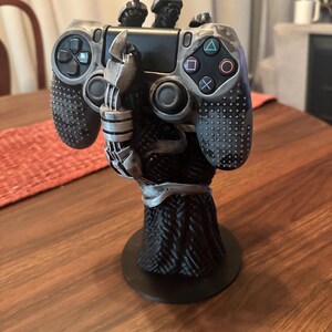 Puede incluir: Un soporte para mando de videojuegos en negro y plata con forma de mano con garras que sostiene un mando con patr&oacute;n de camuflaje gris. El soporte cuenta con una base negra texturizada y un dise&ntilde;o detallado de agarre con aspecto mec&aacute;nico.