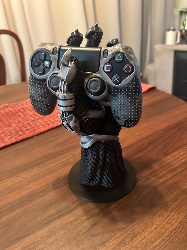 Puede incluir: Un soporte para mando de videojuegos en negro y plata con forma de mano con garras que sostiene un mando con patr&oacute;n de camuflaje gris. El soporte cuenta con una base negra texturizada y un dise&ntilde;o detallado de agarre con aspecto mec&aacute;nico.
