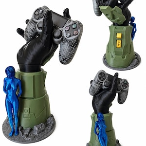 Puede incluir: Un soporte para mando de juegos con una mano negra blindada que emerge de una base cil&iacute;ndrica verde con botones amarillos. Una figura femenina de color azul met&aacute;lico se encuentra junto a la base, sobre una plataforma rocosa de color gris.
