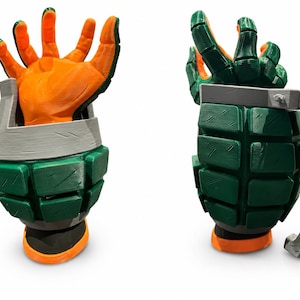 Puede incluir: Una taza impresa en 3D con forma de granada, cuerpo texturizado verde, asa gris y una mano naranja que emerge de la parte superior. El dise&ntilde;o presenta un exterior segmentado con l&iacute;neas de capa visibles y una base plana, en dos &aacute;ngulos.