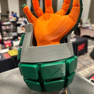 May include: A 3D-printed sculpture of a hand emerging from a grenade-shaped base. The hand is orange with green accents on the fingertips. The base is green and gray, with an orange band at the bottom. The sculpture is likely a fan-made item.