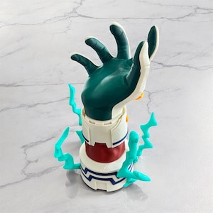 Può includere: Statuetta dettagliata di una mano con palmo e dita verde acqua, dettagli bianchi e una base cilindrica rossa e bianca. Elementi a forma di fulmine verde acqua circondano la base. La mano è in posizione di presa.