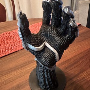 Puede incluir: Un soporte de exhibici&oacute;n con forma de mano esculpida en negro y plata, con un complejo patr&oacute;n de placa de circuito en relieve en la palma y los dedos. La mano est&aacute; en una posici&oacute;n abierta y vertical, montada sobre una base circular negra.