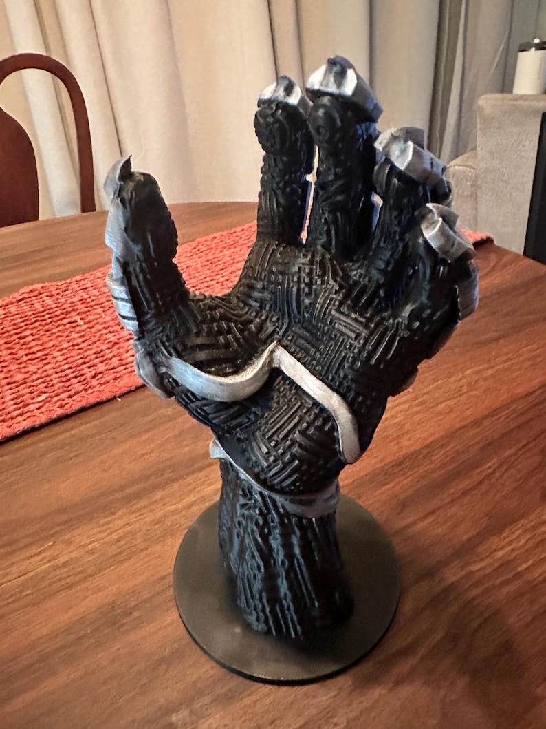 Puede incluir: Un soporte de exhibici&oacute;n con forma de mano esculpida en negro y plata, con un complejo patr&oacute;n de placa de circuito en relieve en la palma y los dedos. La mano est&aacute; en una posici&oacute;n abierta y vertical, montada sobre una base circular negra.