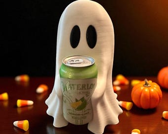 Puede incluir: Decoración de Halloween con forma de fantasma blanco que sostiene una lata verde y plateada de agua con gas Waterloo Lemon-Lime. El fantasma tiene dos ojos ovalados negros. Caramelos de maíz y pequeñas calabazas naranjas están esparcidos.
