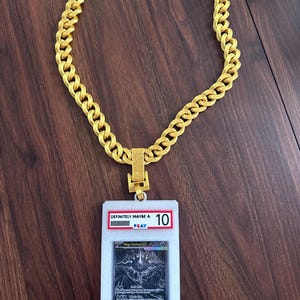 Puede incluir: Un collar de cadena de color dorado con un estuche protector rectangular blanco grande como colgante. El estuche contiene una tarjeta coleccionable y muestra una etiqueta roja en la parte superior con el texto "DEFINITELY MAYBE A 10" y "PSAY" en azul.